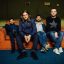 The Black Jackals rock band sitting on a vintage orange sofa, posing for a promotional band photo.